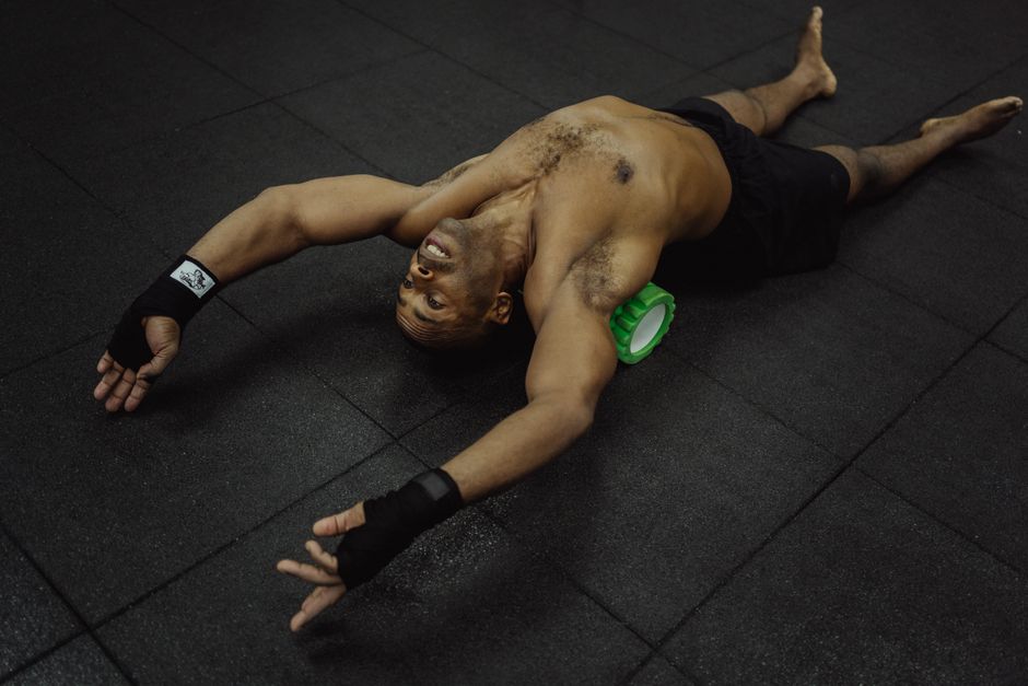 Topless man using foam roller for muscle recovery in gym setting.